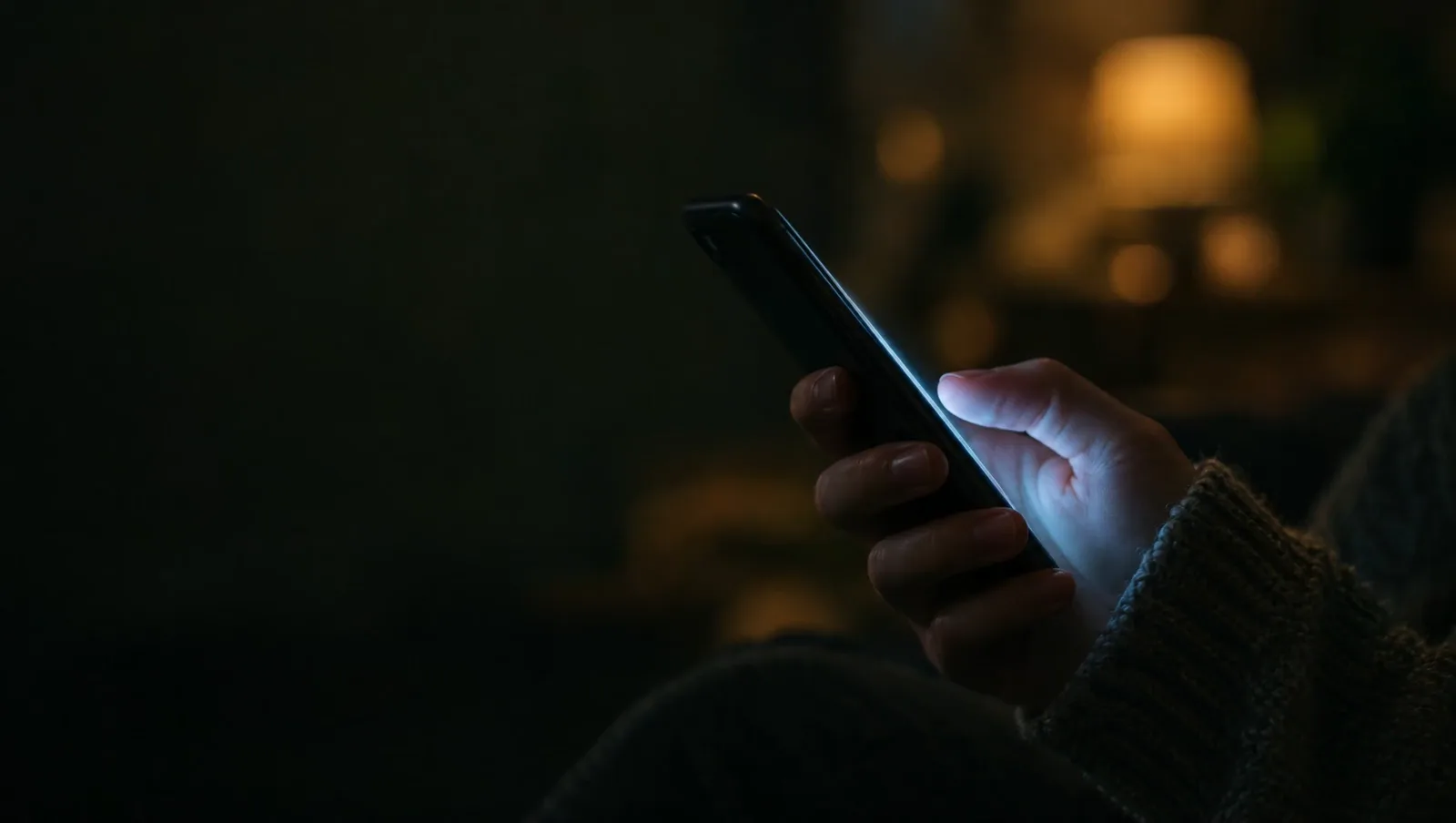 A hand holds a phone in a dim living room, with a warm lamp glowing in the background